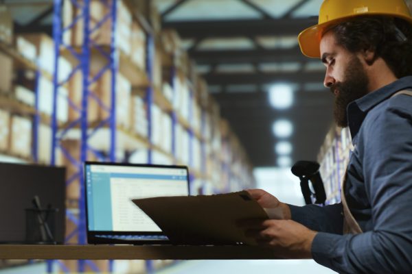 Delivery service associate examines order details on files, working in industrial storage room to ensure shipment in time. Customs compliance staff checks stock tags at import export depot.