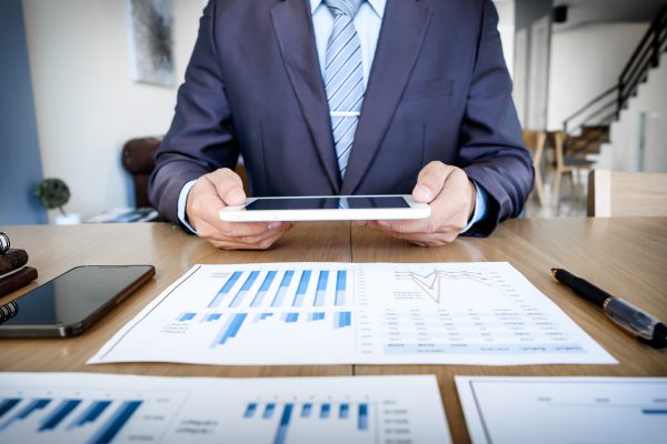 Multitasking. Handsome young man working with touchpad while sitting on the couch in office and financial statistic graph.