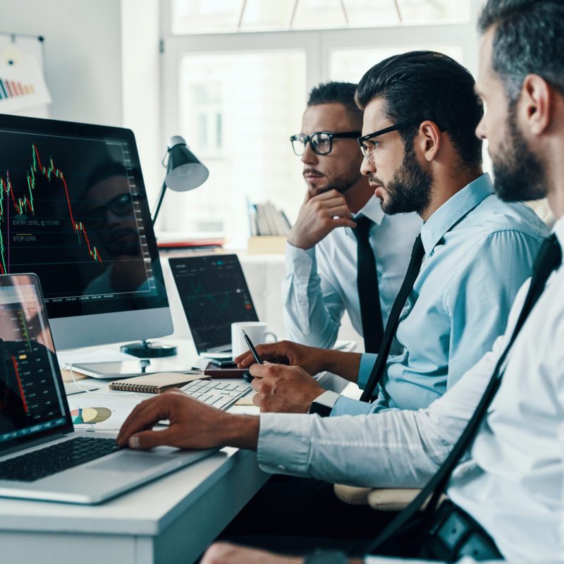 Arbeiten mit Analysen. Eine Gruppe junger, moderner Männer in Anzügen analysiert Börsendaten, während sie im Büro arbeiten.